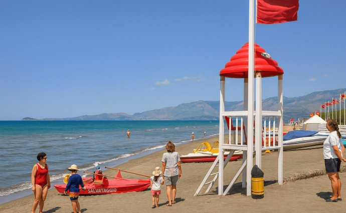 Baia Domizia, spiaggia con torretta di salvataggio, bandiera rossa, pedalò e famiglie in riva.