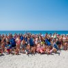 Gruppo di persone sorridenti sulla spiaggia, con il mare azzurro sullo sfondo.