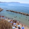 Spiaggia affollata con ombrelloni blu, persone in acqua e scogli sullo sfondo.