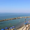 Spiaggia con ombrelloni blu, mare calmo e persone che nuotano; barriera di rocce in lontananza.