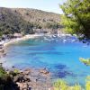 Baia con mare blu, barche ormeggiate e colline verdi. Spiaggia sabbiosa e case in lontananza.